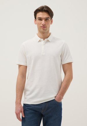 Jonge man die een wit polohemd met korte mouwen en blauwe spijkerbroek draagt, staand met één hand in zijn zak voor een eenvoudige achtergrond.