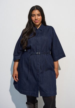 Vrouw met lang donker haar draagt een donkerblauwe denimjas met riem en driekwartmouwen, zwarte panty's en zwarte laarzen tegen een effen achtergrond.
