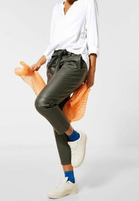 Pantaloni tapered verde oliva con vita regolabile, abbinati a una camicia bianca a maniche lunghe e un wrap arancione testurizzato. Sneakers bianche e calzini blu.