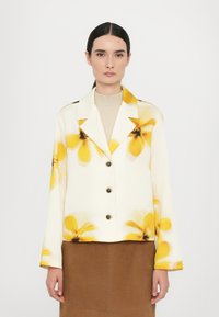 Floral-patterned, cream jacket with yellow accents, notched collar, four black buttons, and long sleeves. Paired with a beige turtleneck and brown skirt.