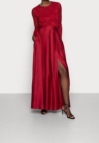 Abito maxi che presenta un corpino in pizzo attillato di un rosso intenso, maniche lunghe e una gonna in satin fluida con uno spacco laterale. Tacchi neri con cinturini completano il look.