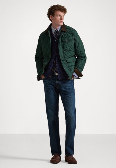 Quiltet grønn jakke med brun kordfløyelskrage, marineblå genser, mønstret slips, jeans og brune sko på en ensfarget bakgrunn.