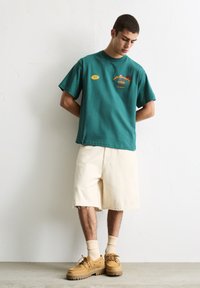 T-shirt en coton teal avec des imprimés graphiques ; shorts en denim crème avec des bords effilochés ; chaussures marron ; chaussettes crème côtelées. Style minimaliste.