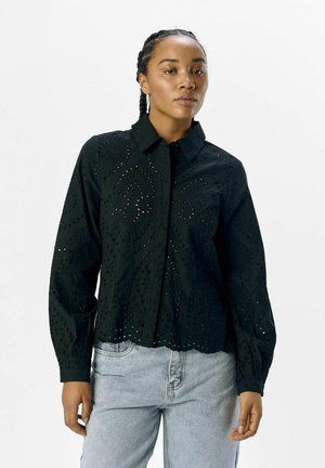 Chemise noire à manches longues, avec un col, des motifs découpés complexes et un ourlet festonné. Conçue en tissu léger.