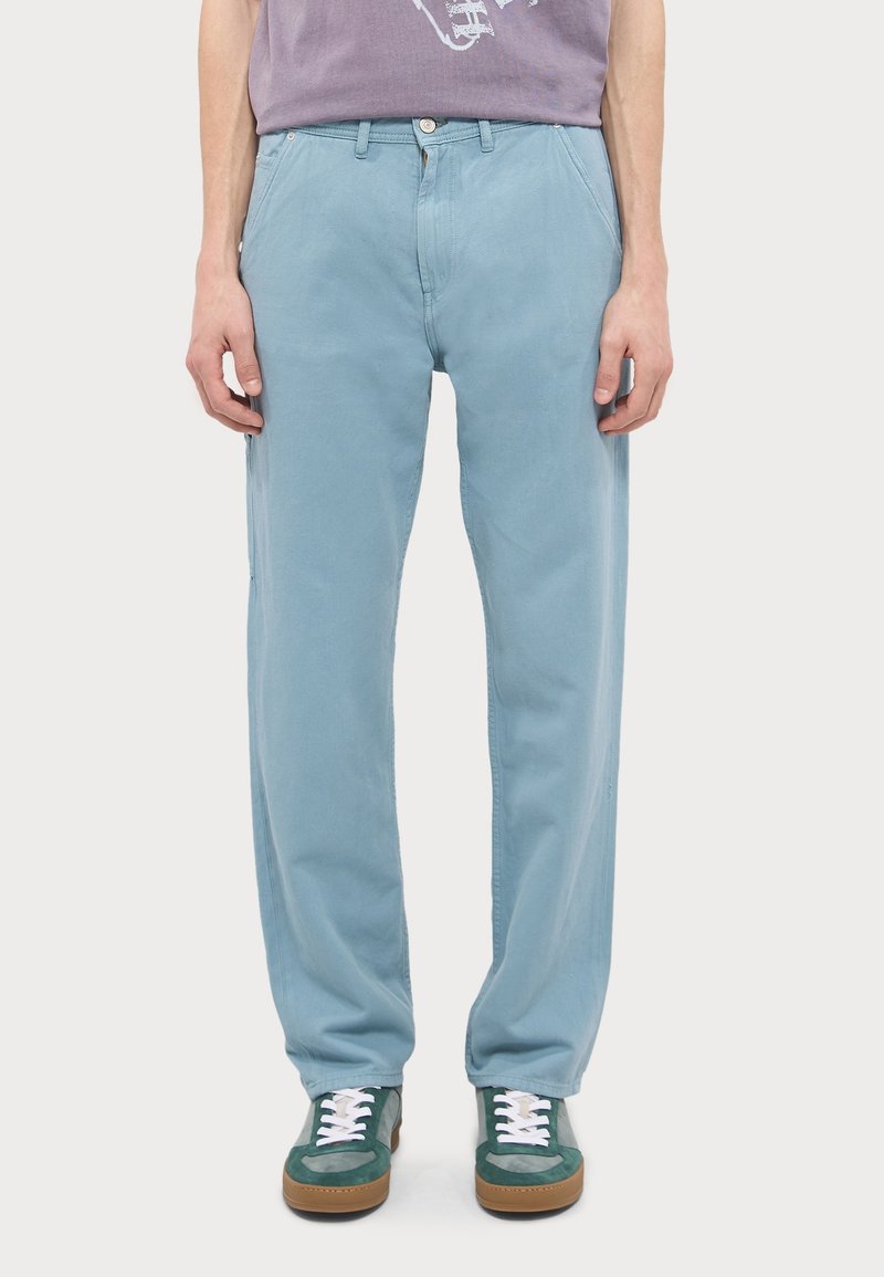 Personne portant un pantalon droit bleu clair et des baskets vertes avec des lacets blancs, debout devant un fond clair uni.