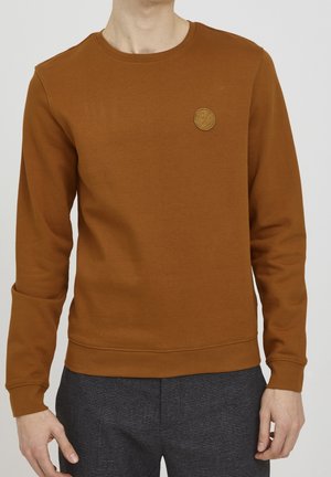 Mann trägt ein rostfarbenes Sweatshirt mit Rundhalsausschnitt und einem kleinen, runden Logo-Patch auf der linken Brust, kombiniert mit dunkelgrauen gemusterten Hosen.