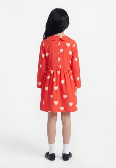 Vaikas su ilgais tamsiais plaukais, vilkintis raudoną suknelę su baltais širdelių raštais, baltomis kojinėmis ir juodais batais, stovintis atsukti nugarą.