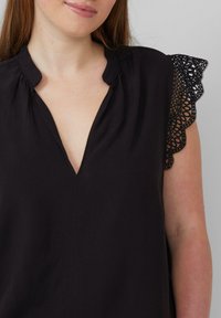 Haut noir avec un col en V et des fronces au niveau du col. Les manches courtes sont ornées de dentelle avec des découpes circulaires, ajoutant une texture au design.