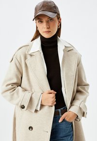 Abrigo de mezcla de lana beige con grandes solapas y botones de doble botonadura, superpuesto a un suéter negro de cuello alto y una camisa blanca, combinado con denim.