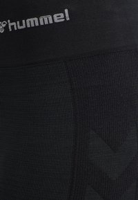 Gros plan sur un tissu noir texturé avec un logo gris "hummel" et un motif chevron discret sur le côté droit.