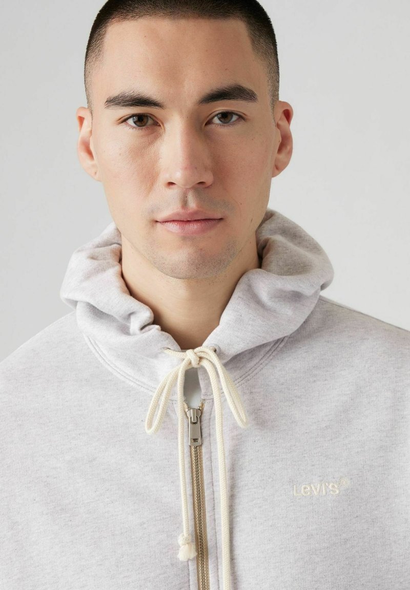 Grauer Kapuzenpullover mit Reißverschluss und Kordelzug, ausgestattet mit einem beigen Reißverschluss und dem bestickten Levi's-Logo. Weicher Stoff mit glatter Textur.
