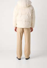 Cremefarvet pufferjakke med hætte, der har et quiltet design. Parret med beige, lige ben bukser og sorte sko. Kun set fra ryggen.