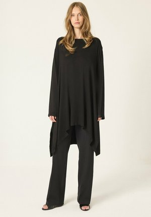 Jeune femme aux longs cheveux blonds portant un haut noir ample à manches longues et un pantalon noir à jambes larges, debout devant un fond uni.
