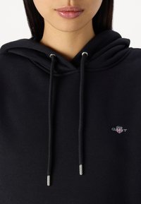 Felpa nera con una texture morbida, dotata di tasca frontale, coulisse con punte argentate e un piccolo logo ricamato sul petto.