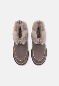 Zapatillas de estar por casa de ante marrón con forro de piel gris, costuras decorativas alrededor de los bordes y detalles en textil con estampado cerca del talón.