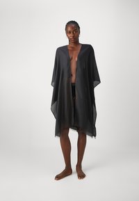 Musta läpikuultava kimono, joka on valmistettu kevyestä kankaasta, avonaisella etuosa, leveillä hihoilla ja tupsullisella helmalla, käytetään uimapuvun päällä.