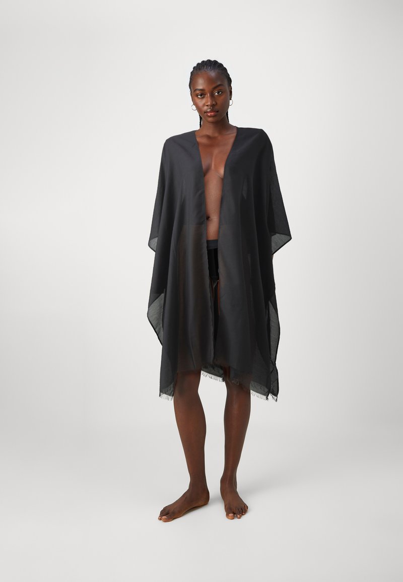Musta läpikuultava kimono, joka on valmistettu kevyestä kankaasta, avonaisella etuosa, leveillä hihoilla ja tupsullisella helmalla, käytetään uimapuvun päällä.