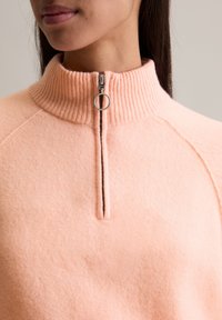 Jasnopomarańczowy polarowy pulower z zamkiem błyskawicznym, z kołnierzem w prążek i subtelnym przeszyciem wzdłuż zamka. Miękka faktura.