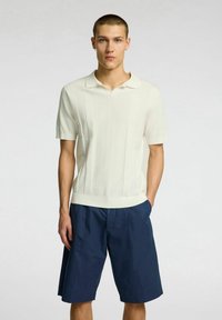 Polo shirt a maniche corte in maglia di colore crema, con righe verticali e colletto piatto. Abbinato a pantaloni corti blu navy su misura.