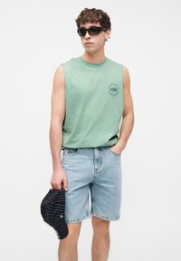 Νεαρός άνδρας με αμάνικο πράσινο πουκάμισο και ανοιχτόχρωμα denim σορτς, κρατώντας ένα μαύρο καπέλο με ρίγες στο ένα χέρι, φοράει μαύρα γυαλιά ηλίου.