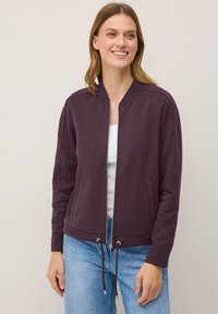 Smiling woman wearing a purple bomber jacket over a white top and blue jeans, standing against a light neutral background.
