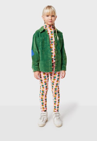 Grüne Cordjacke mit blauen Akzenten, farbenfrohe gemusterte Leggings auf einem pinken Hintergrund und weiße Sneaker.