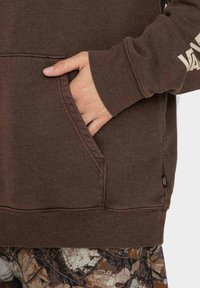 Brązowa bluza z kapturem z przednią kieszenią i ściągaczami na rękawach, z białym logo na rękawie. Tkanina o fakturze i luźny krój.