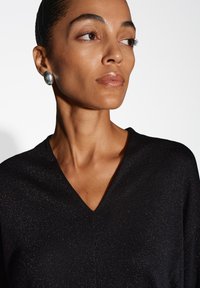 Maglione nero con texture e scollatura a V, caratterizzato da accenti scintillanti. Il modello indossa ampi orecchini argentati riflettenti, mostrando uno stile minimalista.