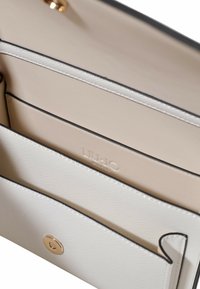 Interno bianco testurizzato di una borsa a tracolla con due scomparti, hardware dorato e un accento nero lungo i bordi. Logo del brand inciso visibile.