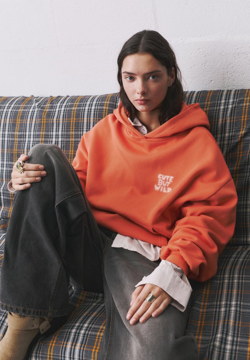 Oversized orange hoodie with "Cute But Wild" embroidery, worn over a white striped shirt, paired with loose gray jeans on a patterned sofa.
