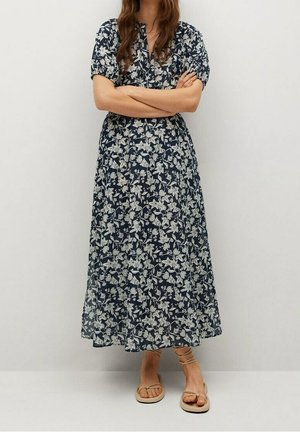 Frau trägt ein dunkelblaues, knöchellanges Kleid mit weißen Blumenmustern und sandfarbenen flachen Sandalen, steht mit verschränkten Armen vor einem schlichten Hintergrund.