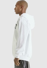 Haut à capuche en coton blanc avec une poche kangourou et un imprimé logo noir. Le design comprend des poignets côtelés et une capuche avec cordon.