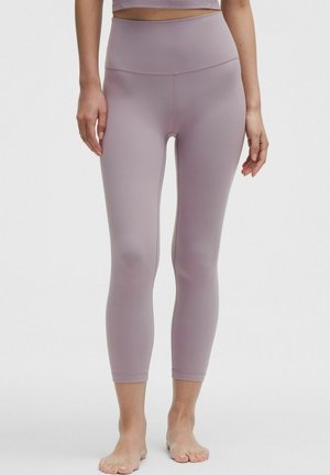 Femme portant des leggings mauves taille haute et un top court assorti, debout pieds nus contre un fond clair uni.