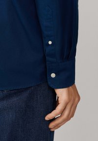 Manica di camicia blu navy con polsino abbottonato, caratterizzata da una superficie in cotone liscia e un dettaglio di design appuntito. Mano posata vicino a un tessuto in denim.