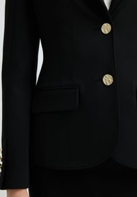 Blazer nero con bottoni dorati, design strutturato con due tasche frontali e texture del tessuto liscia. Vestibilità slanciata e su misura.