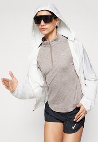 Bijela lagana jakna s kapuljačom, smeša majica kratkih rukava i crne kratke trke. Tamne naočale i vidljivi Nike logotip.
