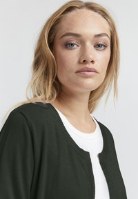Donkergroene cardigan met een ronde halslijn, lichte stof en ribbelranden, gedragen over een witte shirt. Soepele textuur, minimalistisch ontwerp.