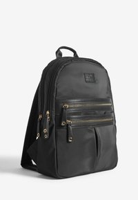Sac à dos en nylon noir avec plusieurs poches zippées, fermetures éclair dorées et une étiquette en cuir texturé. Conçu pour un ajustement ergonomique.