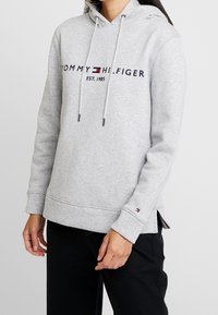 Osoba ubrana w jasnoszary hoodie Tommy Hilfiger z sznurkami i logo na klatce piersiowej, w połączeniu z czarnymi spodniami.