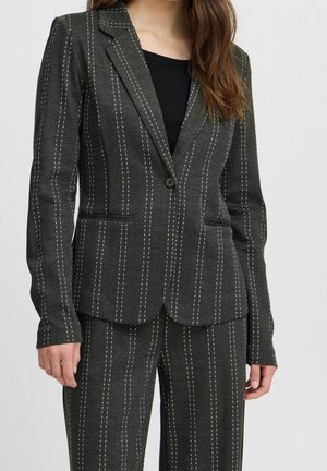 Vrouw die een donkergrijs krijtstreeppakje en bijpassende broek draagt over een zwarte top, staand tegen een effen lichte achtergrond.