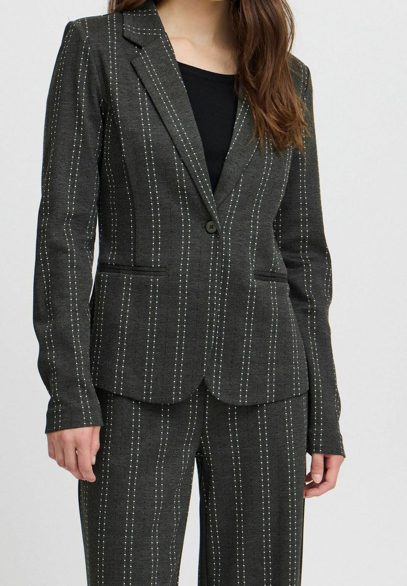 Femme portant une veste de costume gris foncé à fines rayures et un pantalon assorti par-dessus un haut noir, debout devant un fond clair uni.