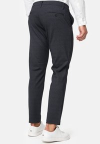 Hombre vistiendo pantalones slim-fit gris oscuro con un bolsillo trasero abotonado y zapatillas blancas, mostrado de espaldas contra un fondo blanco.