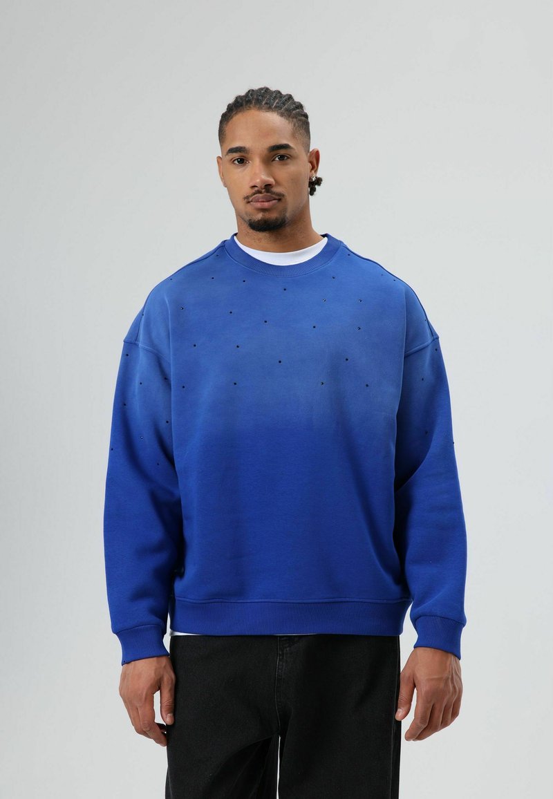 Sweat-shirt bleu avec un design en dégradé, ornée de détails noirs et de poignets côtelés. Porté avec un pantalon noir, le mannequin pose sur un fond clair.
