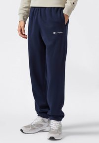 Pantalones de chándal de felpa azul marino con cintura elástica y tobillos ajustados, que presentan un logo bordado en blanco y bolsillos laterales. Combinados con zapatillas grises.