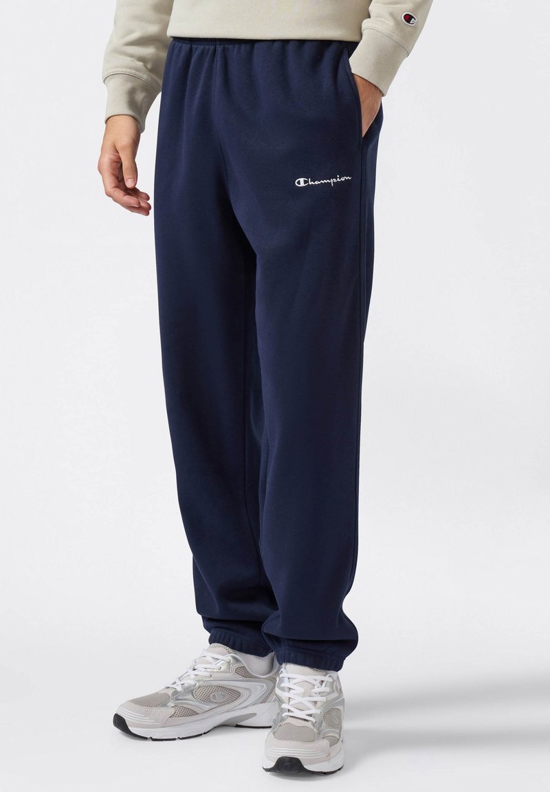 Pantalones de chándal de felpa azul marino con cintura elástica y tobillos ajustados, que presentan un logo bordado en blanco y bolsillos laterales. Combinados con zapatillas grises.