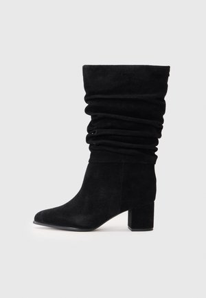Bottes cheville en suède noir avec un design plissé, orteil pointu et un talon épais. La texture est douce et lisse, sans quincaillerie visible.