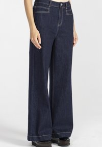 Kobieta w ciemnoniebieskich dżinsach o wysokim stanie i szerokich nogawkach oraz czarnych butach na platformie, stoi na jednolitym białym tle.