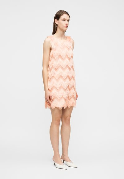 Γυναίκα στέκεται φορώντας αμάνικο μίνι φόρεμα χρώματος ροδάκινου με κρόσσια και σχέδιο ζιγκ-ζαγκ, καθώς και λευκά μυτερά παπούτσια με χαμηλό τακούνι, μπροστά από ένα απλό φόντο.
