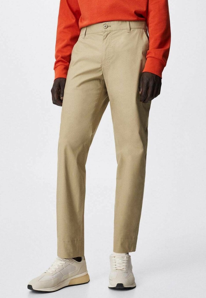 Personne portant un pantalon chino beige droit, des baskets blanches et un haut orange à manches longues, debout devant un fond uni.