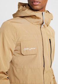 Giacca tan con cappuccio, dotata di chiusura frontale con zip, tasca sul petto con logo bianco e tessuto testurizzato. Include dettagli riflettenti.